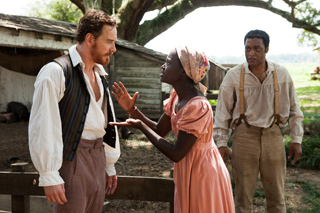 A man in a vest and a woman in a pink dress gesture during a heated discussion near a wooden shack.