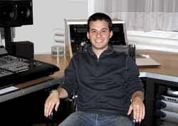 A smiling man in a dark zip-up sweater sits at a desk in a music studio with recording equipment.