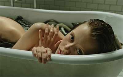 A woman with wet hair lies in a white bathtub, resting her chin near the edge.