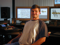 A man in a t-shirt sits in a studio surrounded by computer monitors and audio mixing equipment.
