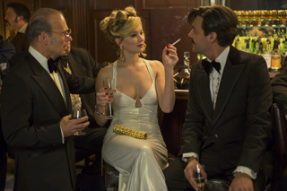 A woman in a white gown smokes a cigarette while talking to two men in tuxedos at a bar.