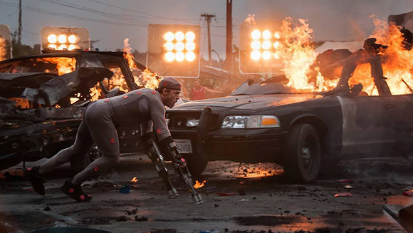 A person in a grey bodysuit with red accents crouches near a black car engulfed in flames.