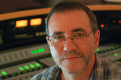 A man with short hair and glasses smiling in a recording studio with audio equipment in the background.