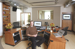 A man sits at a professional recording studio desk surrounded by monitors, mixing consoles, and audio gear.