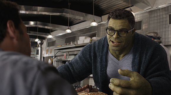 A large green-skinned man with glasses and a blue cardigan sits across from another person.