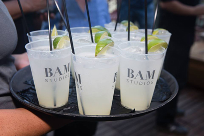 A person holds a black tray filled with several clear plastic cups of pale yellow drinks, each with a lime wedge and black st
