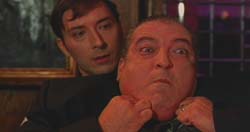A man pulls the cheeks of another man, stretching his face outward.