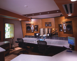 A large mixing console sits in a recording studio with studio monitors and two black office chairs.