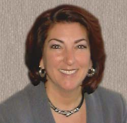 A smiling woman with shoulder-length reddish-brown hair, wearing a grey blazer and a silver necklace.