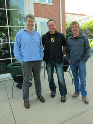 Three men standing side by side on a patio in front of a brick building.