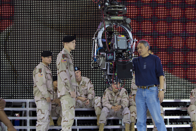 Soldiers in desert camouflage stand near a large camera rig and a man in a blue shirt before an LED screen.