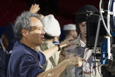 A man in a blue shirt points toward a camera while gesturing to a person in military camouflage.