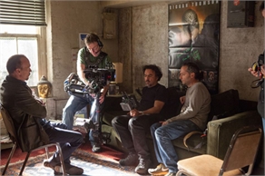 A camera operator films two men sitting on a couch in a rustic indoor setting.
