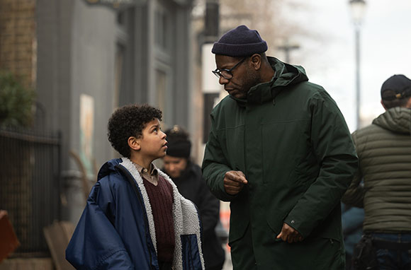 A man in a green jacket and blue beanie leans down to talk to a young boy in a blue jacket on a sidewalk.