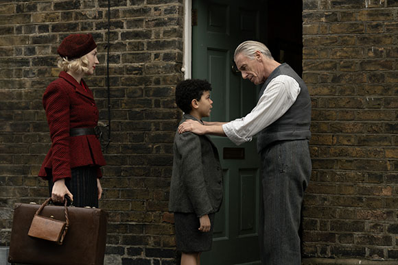A woman in a red coat stands by a suitcase while an older man places his hands on a young boy's shoulders.