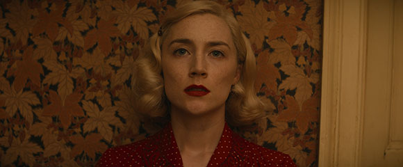 A blonde woman with red lipstick stares directly at the camera against patterned wallpaper.
