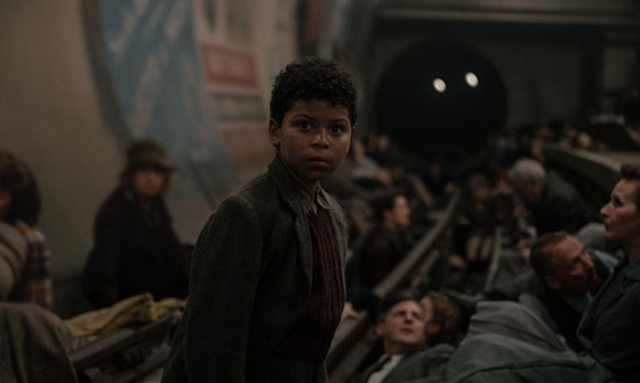 A young boy in a dark jacket looks back while sitting among a crowd in a dimly lit setting.
