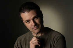 A man in a brown sweater looking toward the camera against a dark, shadowed background.