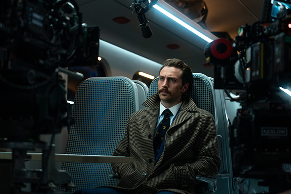 A man with a mustache wearing a brown patterned jacket stands between professional film cameras.