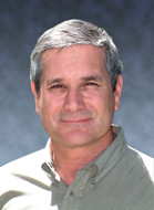 A middle-aged man with short graying hair smiling slightly against a blue background.