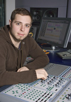 A man in a brown hoodie sits at a large audio mixing desk in a recording studio.