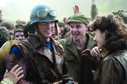 A soldier in a helmet and goggles smiles at a woman in a crowd of cheering people.