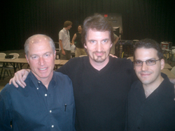 Three men smiling and posing together in a dimly lit room with tables and chairs in the background.