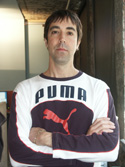 A man with dark hair wearing a white and dark purple Puma long-sleeve shirt with arms crossed.