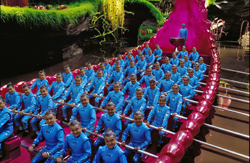 A large group of people in matching blue uniforms seated in a long, pink, boat-shaped structure.