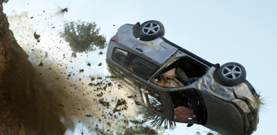 A silver SUV flips through the air amidst flying dirt and debris against a pale sky.
