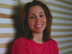 A smiling woman with shoulder-length brown hair wearing a pink top against a beige and white striped wall.