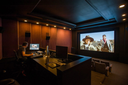 A person sits at a desk with monitors in a dark room facing a large movie screen playing a film.