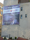 A large rectangular billboard mounted on a light-colored building wall with text and logos.