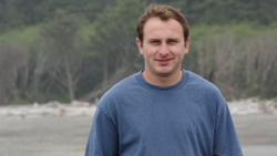 A man with short brown hair wearing a blue t-shirt stands in front of a blurred forest background.