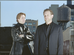 Two men in dark clothing pose on a rooftop against a city skyline and water tower.