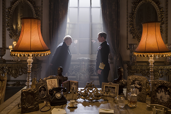 Two men stand in a grand room with ornate lamps and a desk covered in photographs and a rotary phone. Sunlight streams throug