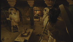 Two large angel statues stand in a dark, arched stone chamber with pillars and stone altars.