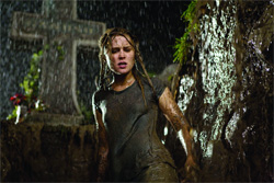 A woman with wet hair and a soaked shirt stands in the rain next to a stone cross and a rocky wall.