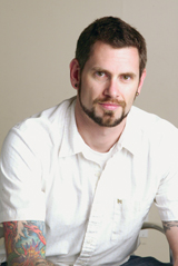 A man with short dark hair, a goatee, and colorful arm tattoos wearing a white button-down shirt.