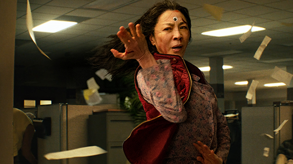A woman with a third eye on her forehead reaches toward the camera in a messy office with flying papers.