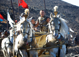 A man in golden armor drives a two-horse chariot across a rocky landscape.