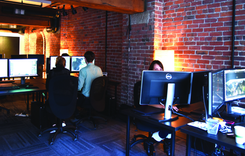 People sit at desks with multiple computer monitors in a dimly lit office with brick walls.