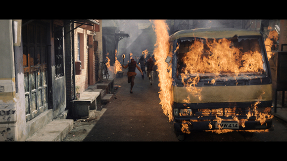 A yellow van is on fire in a narrow, smoky alleyway as people run in the background.