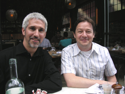 Two men smiling while sitting at a white table in an outdoor dining area.