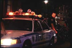 A white NYPD police car with amber emergency lights flashing on its roof at night.