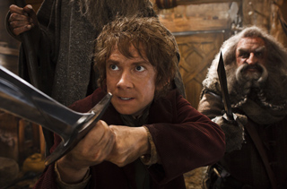 A man with curly hair holds a large sword in a dark, rustic room next to a bearded man.