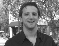 A smiling man in a dark collared shirt standing in front of trees and buildings.