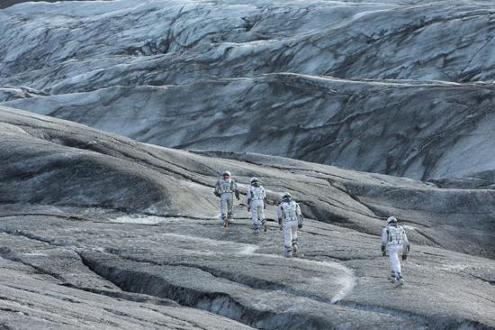 Four people in hiking gear walking across a large, grey, undulating glacier landscape.