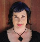 A woman with dark, wavy hair with purple highlights, wearing a black top and a dark pendant necklace.