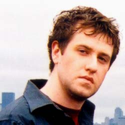 A young man with wavy brown hair looking forward against a pale sky background.